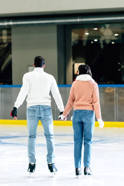 Vista trasera de pareja joven cogida de la mano en la pista de patinaje - foto de stock