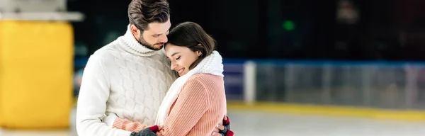 Tiro panorâmico de belo jovem casal em suéteres abraçando na pista de patinação — Fotografia de Stock