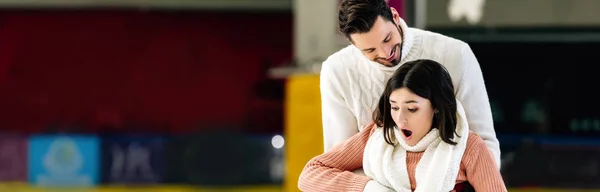 Panoramic shot of handsome smiling man catching scared falling woman on skating rink — Stock Photo