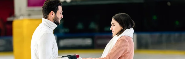 Panoramic shot of beautiful cheerful young couple holding hands on skating rink — Stock Photo