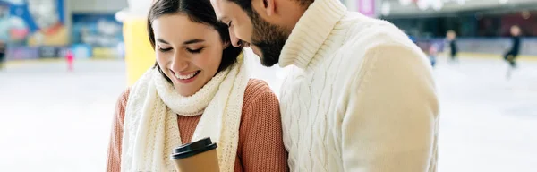 Panoramic shot of beautiful smiling couple holding coffee to go on skating rink — Stock Photo