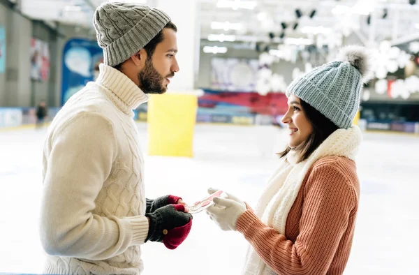 Schöner Mann übergibt Glückwunschkarte am Valentinstag an fröhliche Frau auf der Eisbahn — Stockfoto