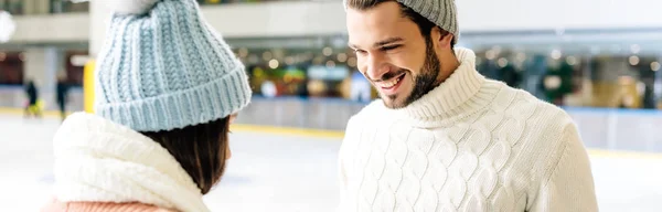 Panoramic shot of young cheerful couple in hats looking at each other on skating rink — Stock Photo