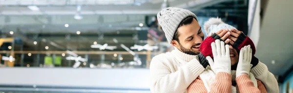 Panoramic shot of handsome cheerful man closing eyes to woman to make a surprise on skating rink — Stock Photo
