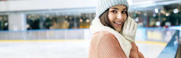 Colpo panoramico di allegra ragazza attraente in maglione, sciarpa, guanti e cappello in piedi sulla pista di pattinaggio — Foto stock