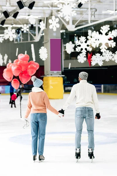 Vista posterior de la pareja con globos rojos en forma de corazón tomados de la mano en la pista de patinaje en el día de San Valentín - foto de stock