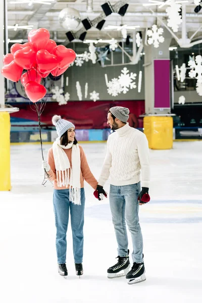 Casal alegre com balões em forma de coração vermelho de mãos dadas na pista de patinação no dia dos namorados — Fotografia de Stock