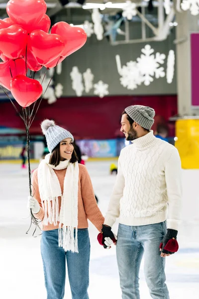 Coppia felice con palloncini rossi a forma di cuore che si tiene per mano sulla pista di pattinaggio il giorno di San Valentino — Foto stock