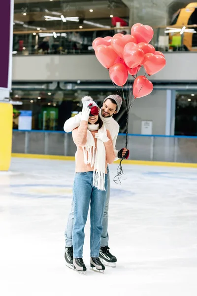 Uomo con palloncini rossi a forma di cuore che chiudono gli occhi alla ragazza sulla pista di pattinaggio il giorno di San Valentino — Foto stock