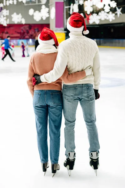 Back view of happy couple in santa hats hugging on skating rink — Stock Photo