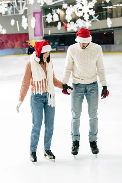 Happy couple in santa hats holding hands while skating on rink — Stock Photo