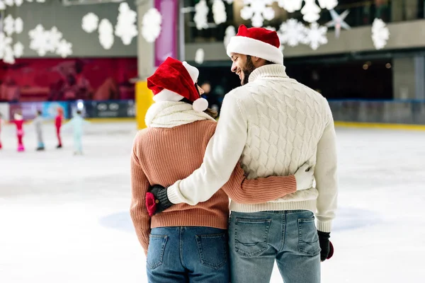 Rückansicht eines glücklichen Paares in Weihnachtsmannmützen, das sich auf der Eisbahn umarmt und anschaut — Stockfoto