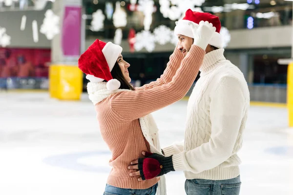 Alegre pareja en santa sombreros pasar la navidad en pista de patinaje - foto de stock