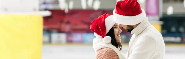 Tiro panorâmico de belo casal feliz em chapéus santa passar Natal na pista de patinação — Fotografia de Stock