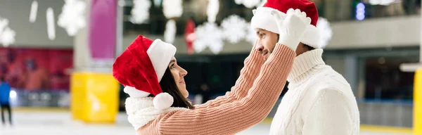 Colpo panoramico di bella coppia felice in cappelli di Babbo Natale trascorrere del tempo insieme sulla pista di pattinaggio — Foto stock