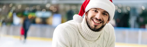 Panoramic shot of smiling man in sweater and santa hat on skating rink — Stock Photo