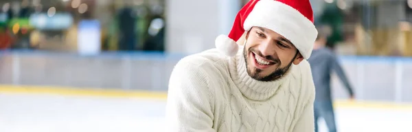 Tiro panorâmico de homem sorridente barbudo em suéter branco e chapéu de santa na pista de patinação — Fotografia de Stock