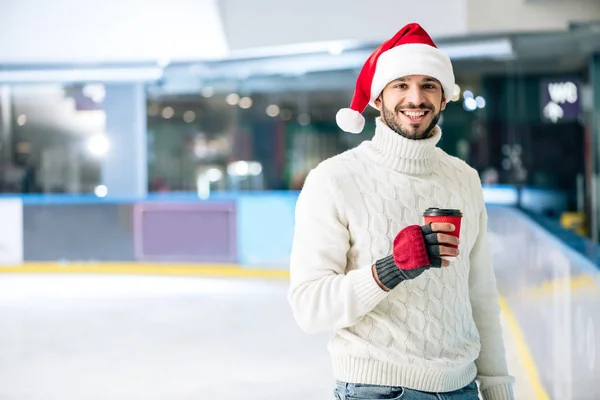 Cheerful man in sweater and santa hat holding coffee to go on skating rink — Stock Photo