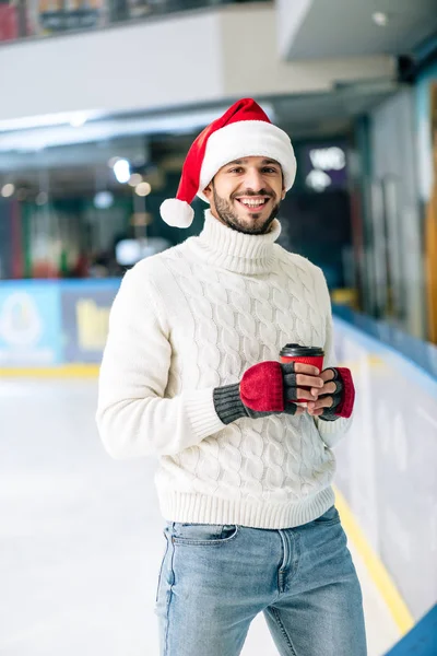 Bel homme joyeux en pull et santa chapeau tenant café pour aller sur patinoire — Photo de stock