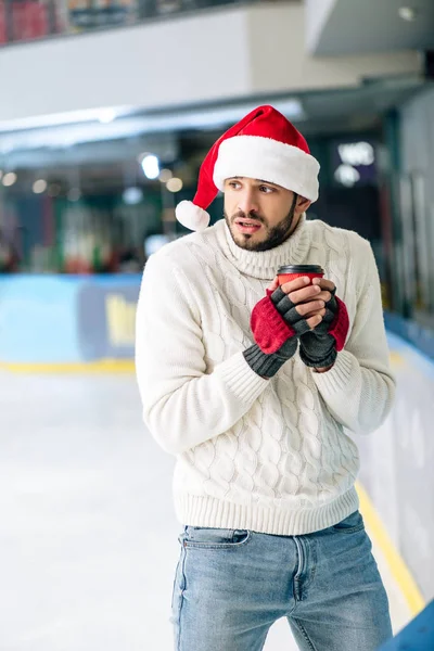 Homme froid en pull et santa chapeau tenant café pour aller sur patinoire — Photo de stock