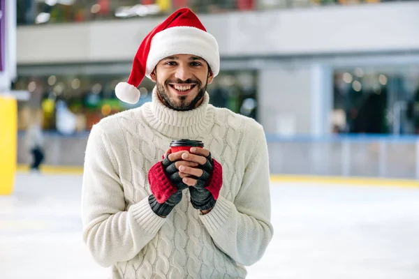 Bell'uomo allegro in cappello di Babbo Natale che tiene il caffè per andare sulla pista di pattinaggio — Foto stock