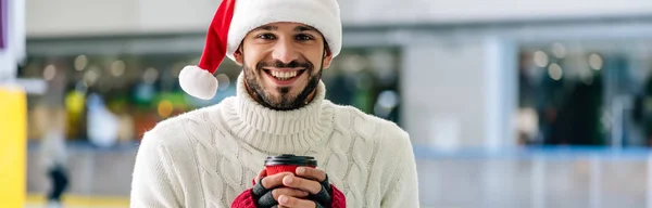 Panoramaaufnahme eines lächelnden Mannes mit Weihnachtsmannmütze, der Kaffee hält, um auf die Eisbahn zu gehen — Stockfoto