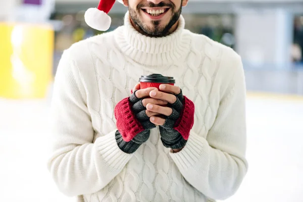 Vista cortada de homem alegre em santa chapéu segurando café para ir na pista de patinação — Fotografia de Stock