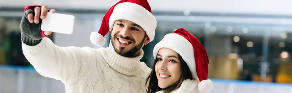 Panoramic shot of happy couple in santa hats taking selfie on smartphone on skating rink — Stock Photo