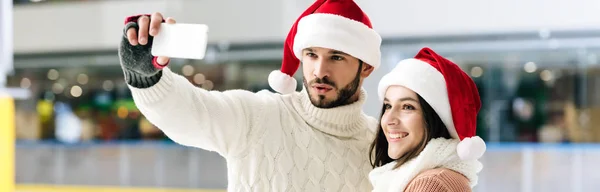 Panoramic shot of beautiful couple in santa hats taking selfie on smartphone on skating rink — Stock Photo