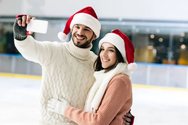 Smiling couple in santa hats taking selfie on smartphone on skating rink at christmastime — Stock Photo