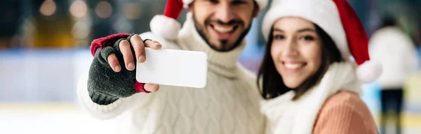 Panoramic shot of smiling couple in santa hats taking selfie on smartphone on skating rink at christmastime — Stock Photo