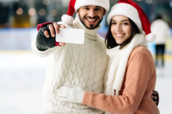 Selective focus of happy couple in santa hats taking selfie on smartphone on skating rink at christmastime — Stock Photo