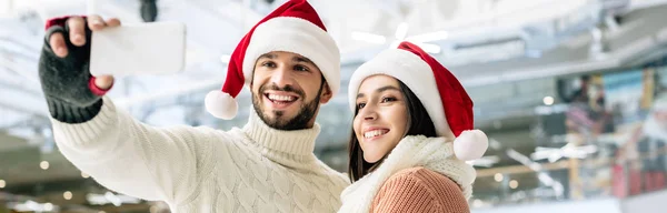 Panoramic shot of happy couple in santa hats taking selfie on smartphone on skating rink at christmastime — Stock Photo