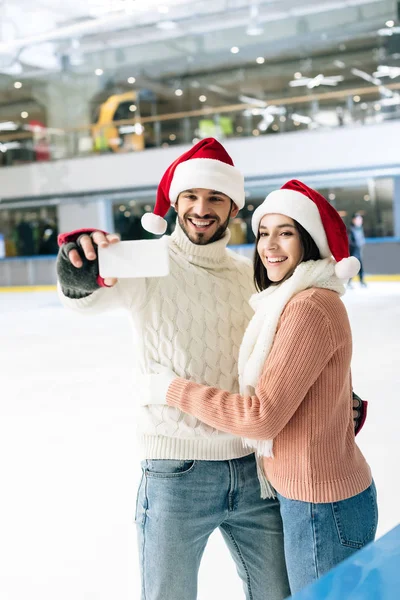 Beautiful happy couple in santa hats taking selfie on smartphone on skating rink at christmastime — Stock Photo