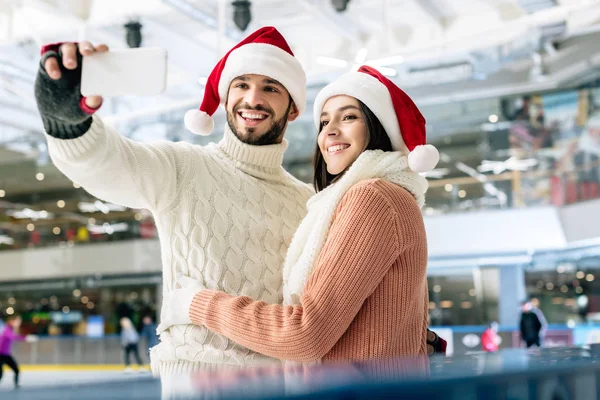 Belo casal alegre em chapéus de santa tirar selfie no smartphone na pista de patinação no Natal — Fotografia de Stock