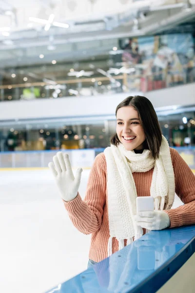 Menina alegre segurando smartphone e mão acenando na pista de patinação — Fotografia de Stock