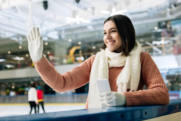 Beautiful smiling girl holding smartphone and waving hand on skating rink — Stock Photo