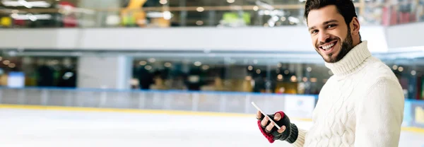 Panoramaaufnahme eines gut aussehenden, lächelnden Mannes mit Smartphone auf der Eisbahn — Stockfoto