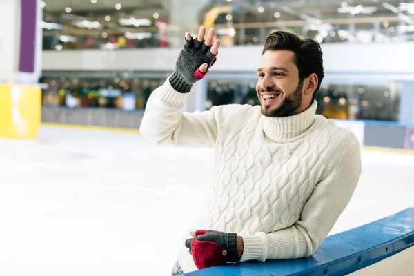 Sonriente hombre en suéter y guantes agitando la mano en la pista de patinaje - foto de stock