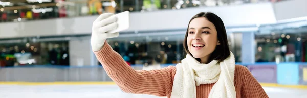 Панорамный снимок привлекательной веселой девушки, делающей селфи на смартфоне на катке — стоковое фото