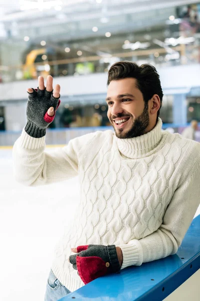 Alegre guapo hombre en suéter y guantes agitando la mano en pista de patinaje - foto de stock