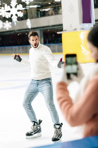 Enfoque selectivo de la mujer tomando la foto del hombre emocionado en el teléfono inteligente en la pista de patinaje - foto de stock