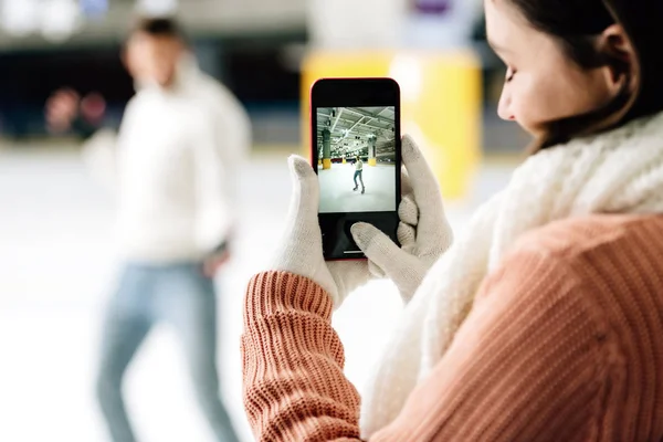 Selective focus of beautiful woman taking photo of man on smartphone on skating rink — Stock Photo