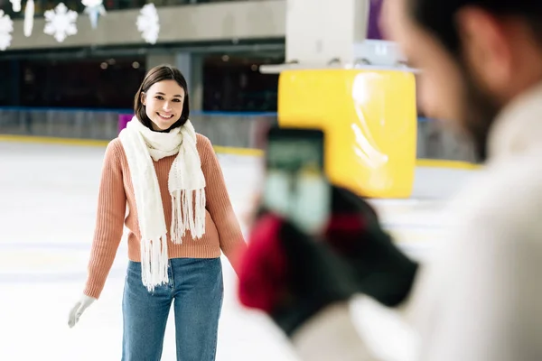 Selective focus of boyfriend taking photo of smiling girlfriend on smartphone on skating rink — Stock Photo