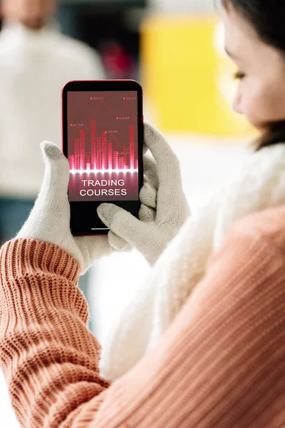 Cropped view of woman in gloves holding smartphone with trading courses app on screen — Stock Photo