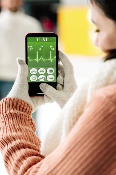 КИЕВ, Украина - 15 ноября 2019 года: обрезанный вид женщины в перчатках, держащей смартфон с приложением для здоровья на экране — стоковое фото