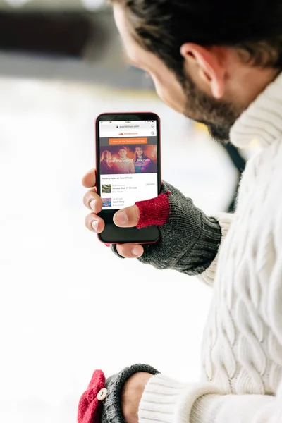 KYIV, UCRANIA - 15 DE NOVIEMBRE DE 2019: vista recortada del hombre con guantes que sostiene el teléfono inteligente con la aplicación soundcloud en la pantalla, en la pista de patinaje - foto de stock