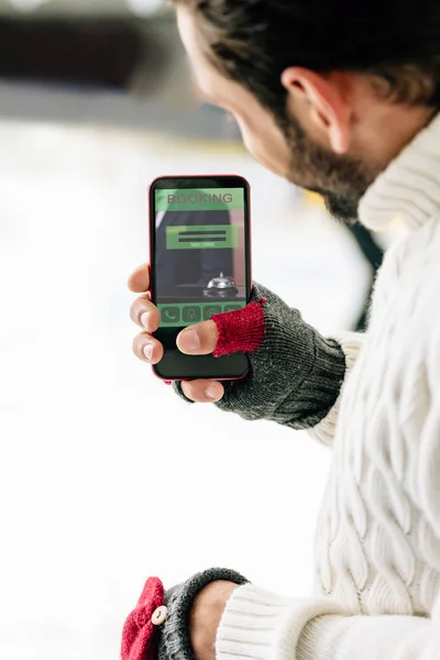 KYIV, UCRANIA - 15 de noviembre de 2019: vista recortada del hombre con guantes que sostiene el teléfono inteligente con la aplicación de reserva en la pantalla, en la pista de patinaje - foto de stock