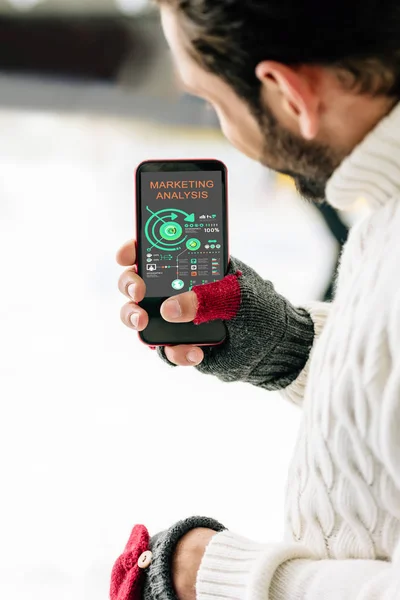 KYIV, UCRANIA - 15 de noviembre de 2019: vista recortada del hombre con guantes que sostiene el teléfono inteligente con la aplicación de análisis de marketing en la pantalla, en la pista de patinaje - foto de stock