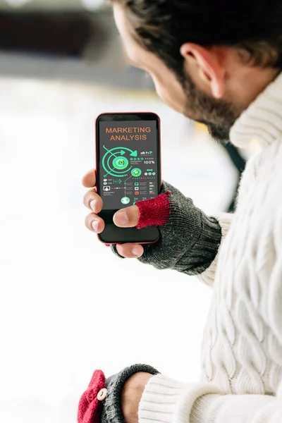 KYIV, UKRAINE - 15 NOVEMBRE 2019 : vue recadrée de l'homme en gants tenant un smartphone avec une application d'analyse marketing à l'écran, sur une patinoire — Photo de stock